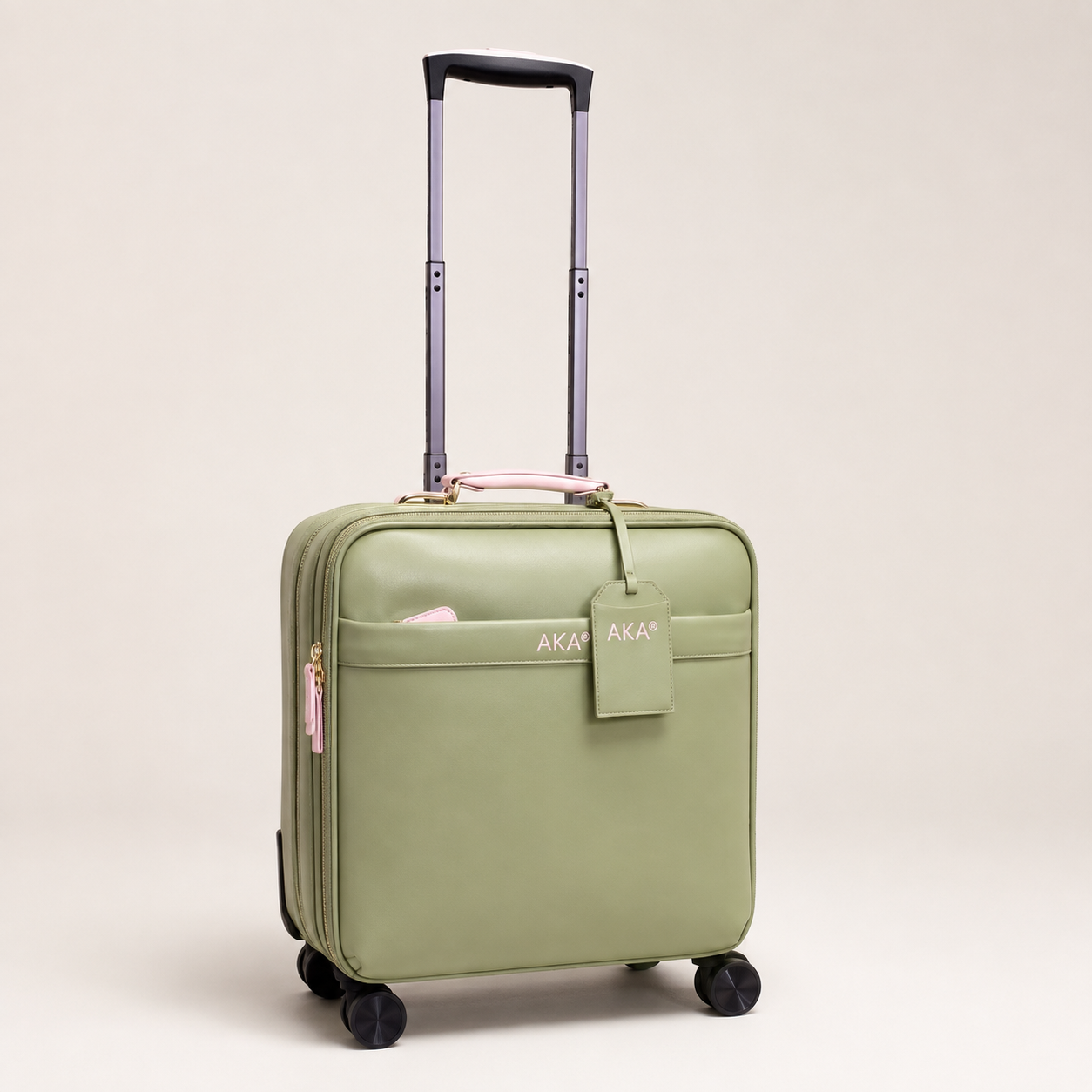 A sage green hard-shell carry-on suitcase with an extended telescoping handle, four black spinner wheels, a front zip pocket, and a matching luggage tag. The brand name 'AKA' is printed in white on the front pocket and the luggage tag.