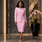 A woman models a light pink two-piece suit with textured fabric and silver crystal embellishments on the jacket, paired with a matching pencil skirt, in a modern lobby setting.